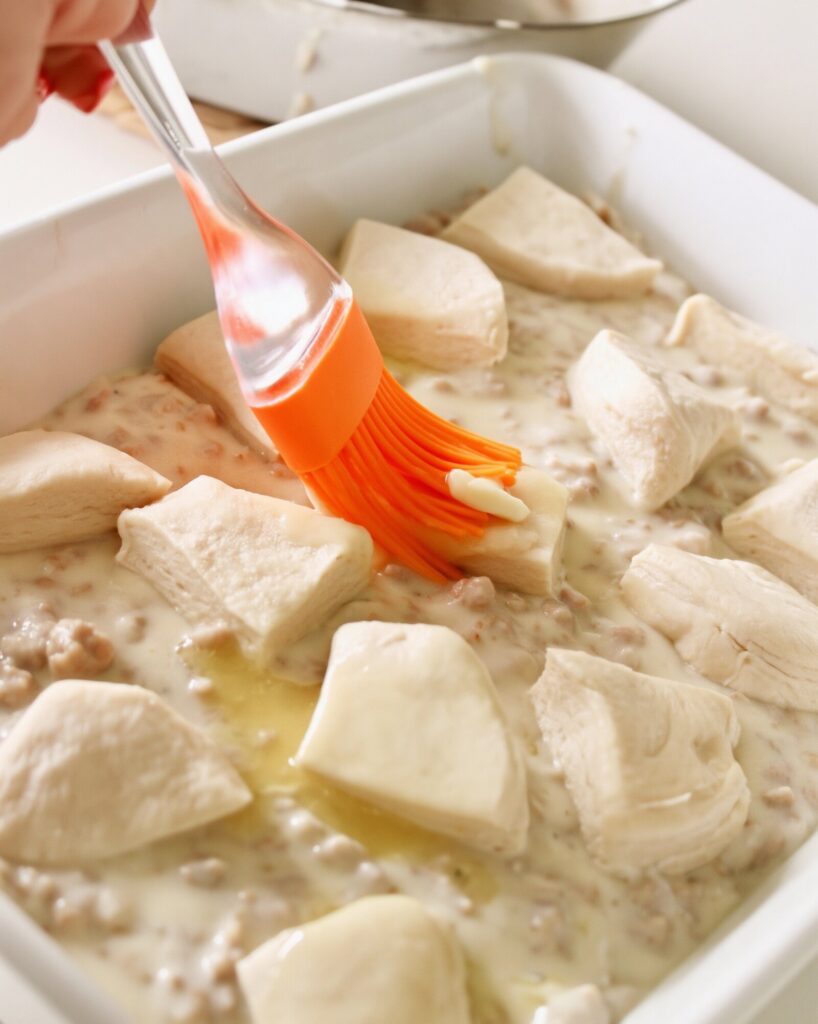 An orange silicone brush spreading melted butter over raw biscuit pieces on sausage gravy in a white baking dish