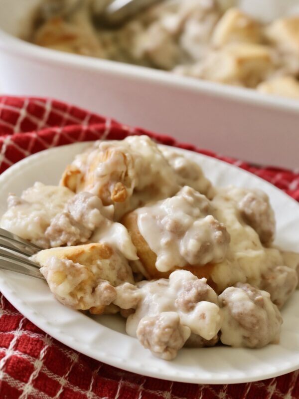 A serving of biscuits and sausage gravy casserole on a white plate with a fork, on a red checked towel, the baking dish behind