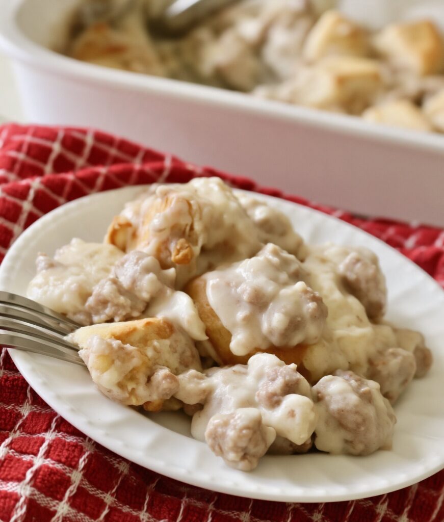A serving of biscuits and sausage gravy casserole on a white plate with a fork, on a red checked towel, the baking dish behind