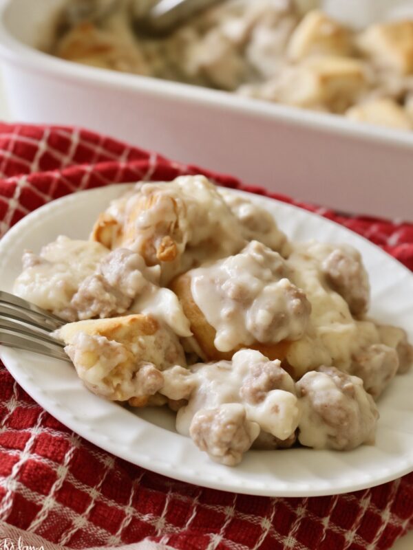 A serving of biscuits and sausage gravy casserole on a white plate with a fork, on a red checked towel, the baking dish behind