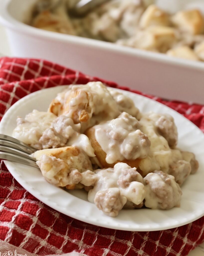 A serving of biscuits and sausage gravy casserole on a white plate with a fork, on a red checked towel, the baking dish behind