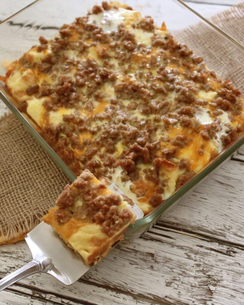 A metal spatula lifting a corner square of sausage breakfast casserole out of a clear glass baking dish set on burlap over whitewashed wood boards