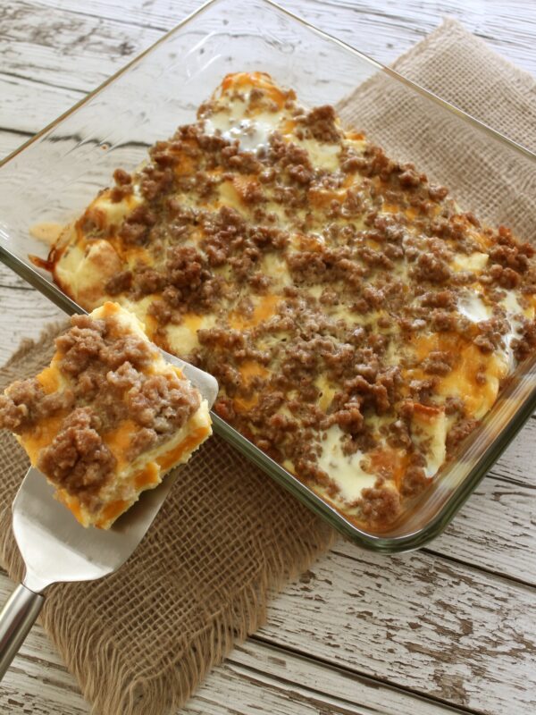 A square of breakfast casserole with sausage crumbles, cheddar and set egg held up on a spatula beside the glass baking dish