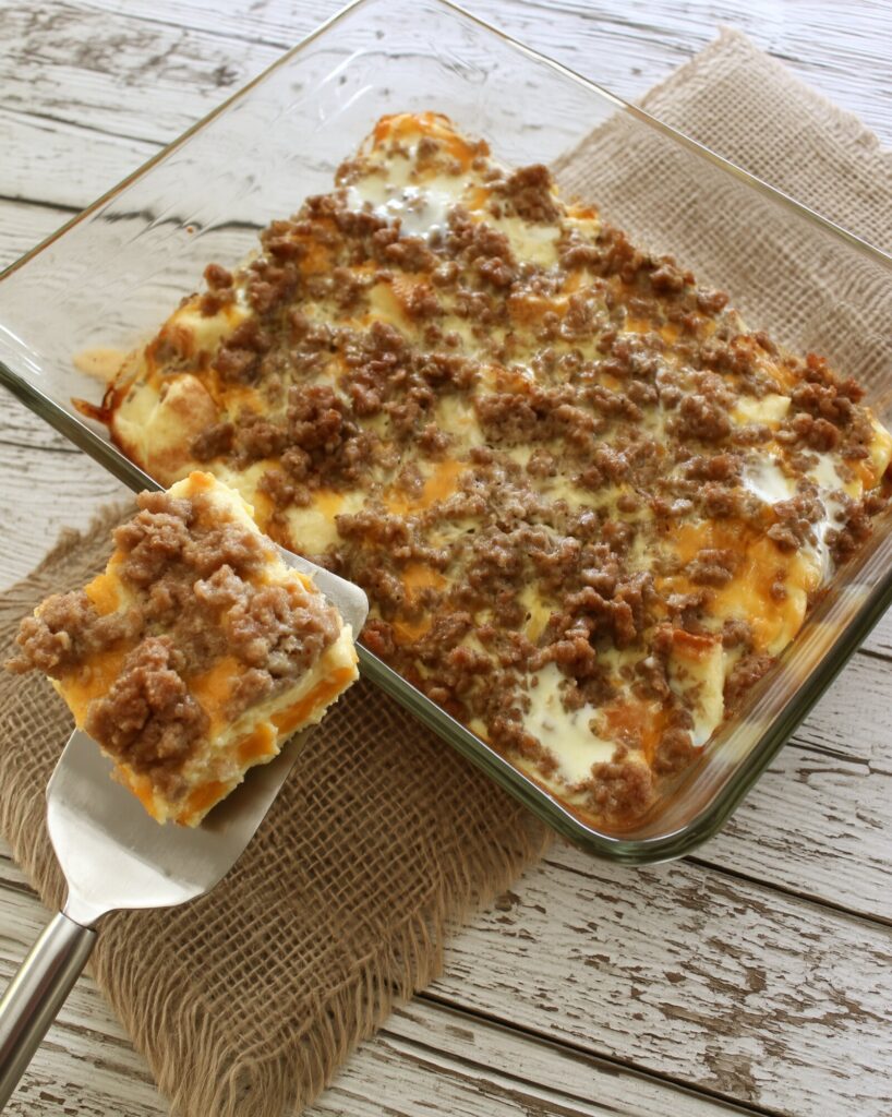 A square of breakfast casserole with sausage crumbles, cheddar and set egg held up on a spatula beside the glass baking dish