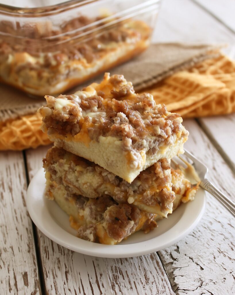 Three squares of sausage breakfast casserole stacked on a small white plate with a fork, the glass baking dish and an orange towel behind on white wood