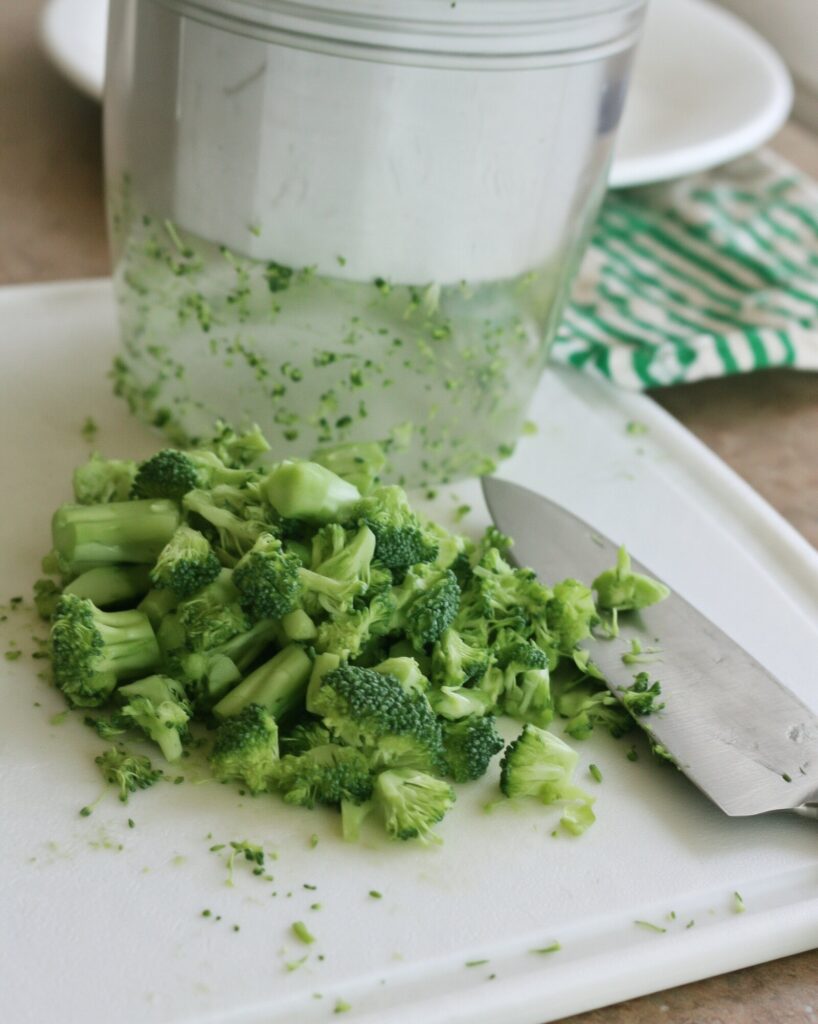 Hand-chopped broccoli in small uneven pieces on a white cutting board next to a chef's knife and a food processor bowl of finely chopped broccoli