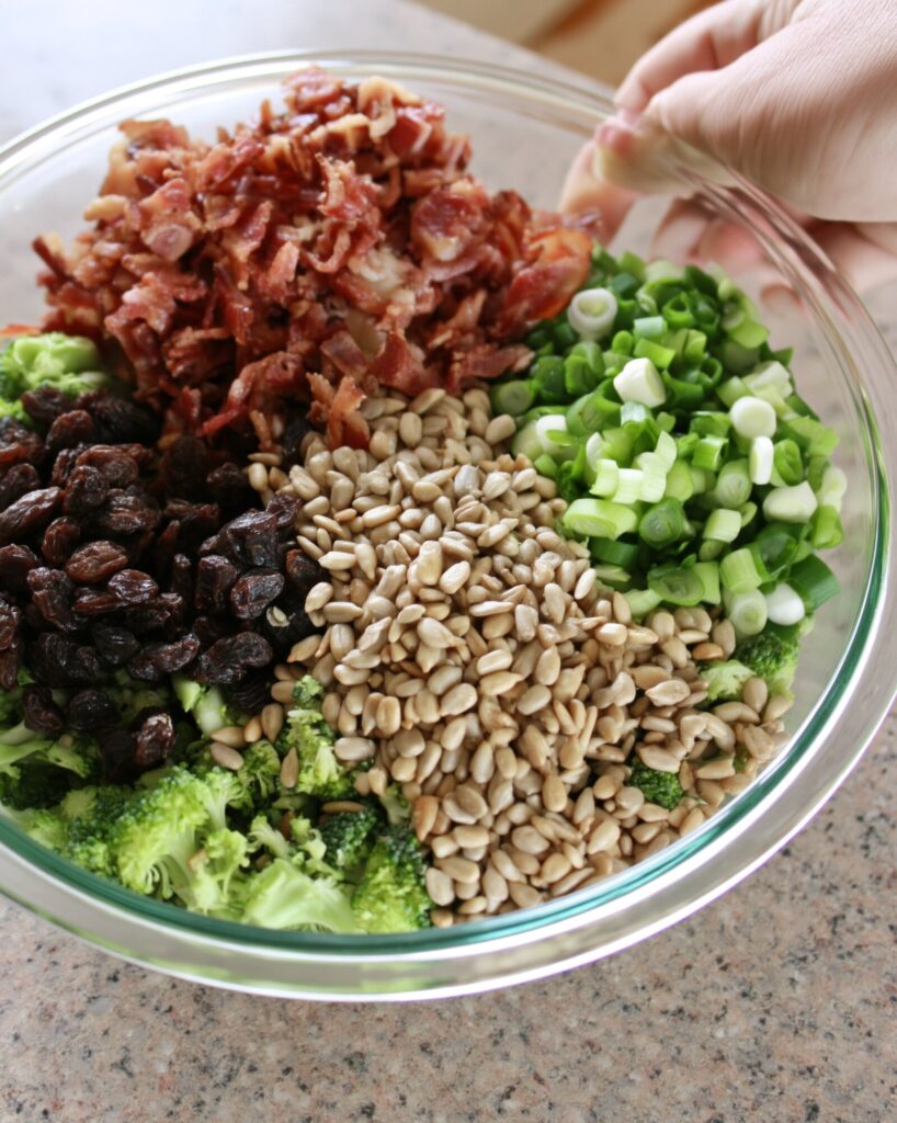 A glass bowl of broccoli topped with crumbled bacon, raisins, sunflower seeds and sliced green onions, a hand tilting the bowl