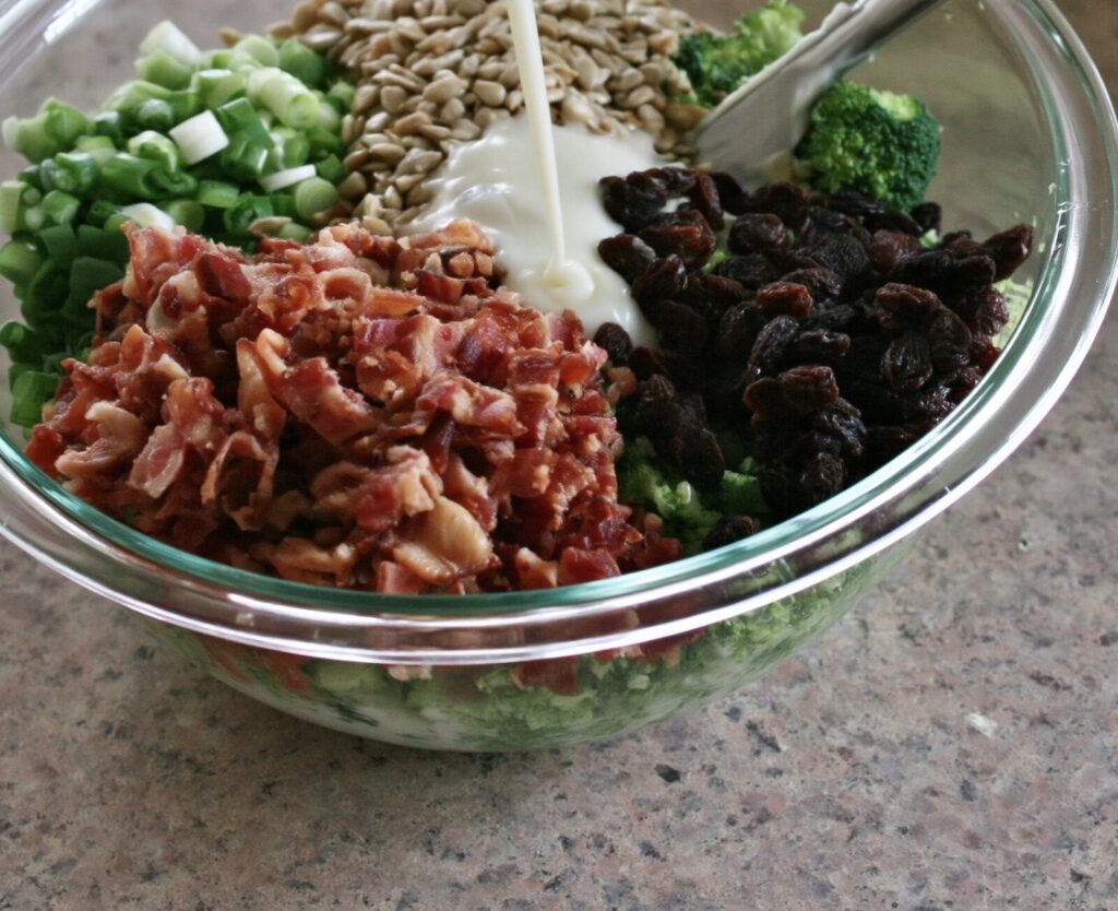 Creamy white dressing pouring into a glass bowl of broccoli, bacon, raisins, sunflower seeds and green onions, a spoon resting on the rim