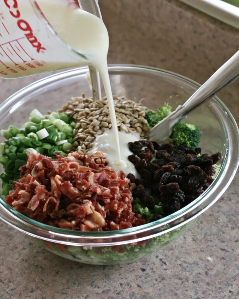 Creamy white dressing pouring from a measuring cup into a glass bowl of broccoli, bacon, raisins, sunflower seeds and green onions, a spoon resting on the rim
