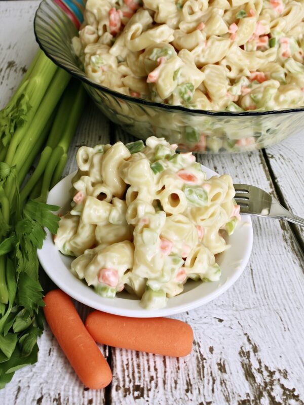 A small white bowl of creamy macaroni salad with a fork resting on the rim, a big glass bowl of salad, celery stalks and whole carrots on whitewashed wood behind it