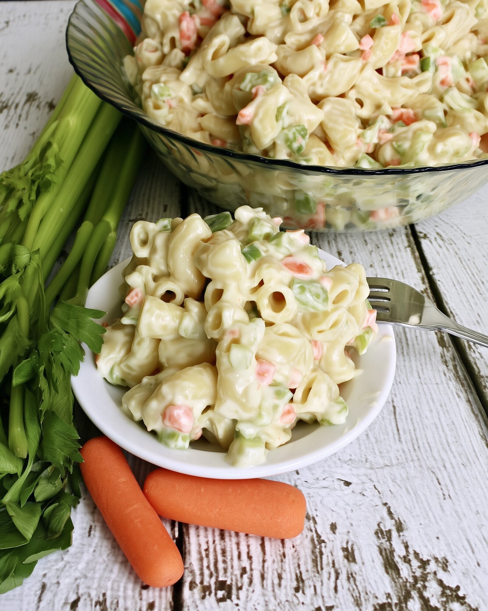 A small white bowl of creamy macaroni salad with a fork resting on the rim, a big glass bowl of salad, celery stalks and whole carrots on whitewashed wood behind it