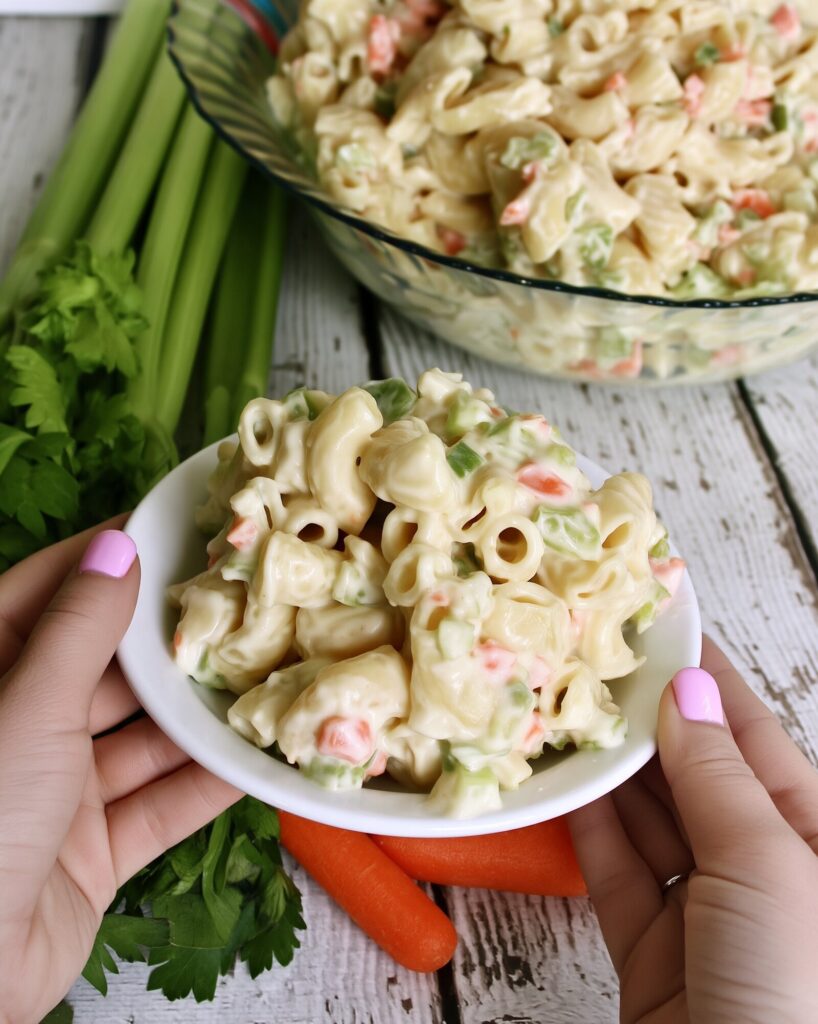 Two hands holding a small white bowl of creamy elbow macaroni salad with bits of carrot and celery, over whitewashed wood with celery and carrots