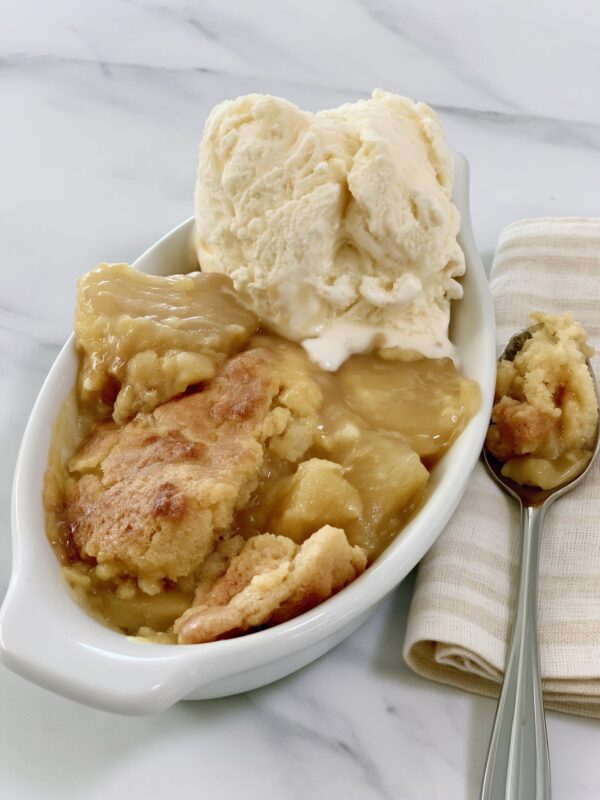 An oval white dish of crock pot apple crisp topped with a scoop of vanilla ice cream, a spoonful resting on a cloth napkin beside it on marble