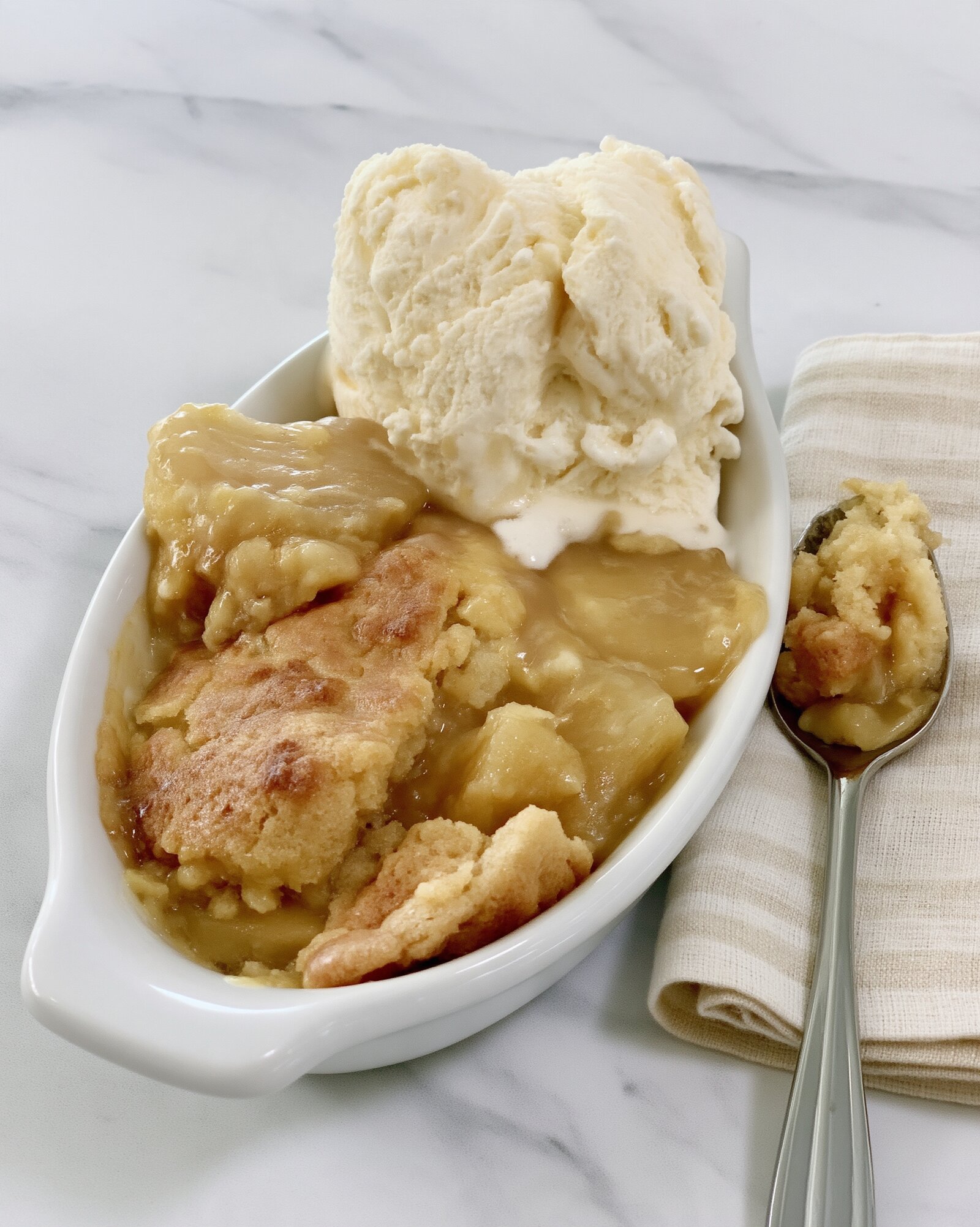 An oval white dish of crock pot apple crisp topped with a scoop of vanilla ice cream, a spoonful resting on a cloth napkin beside it on marble