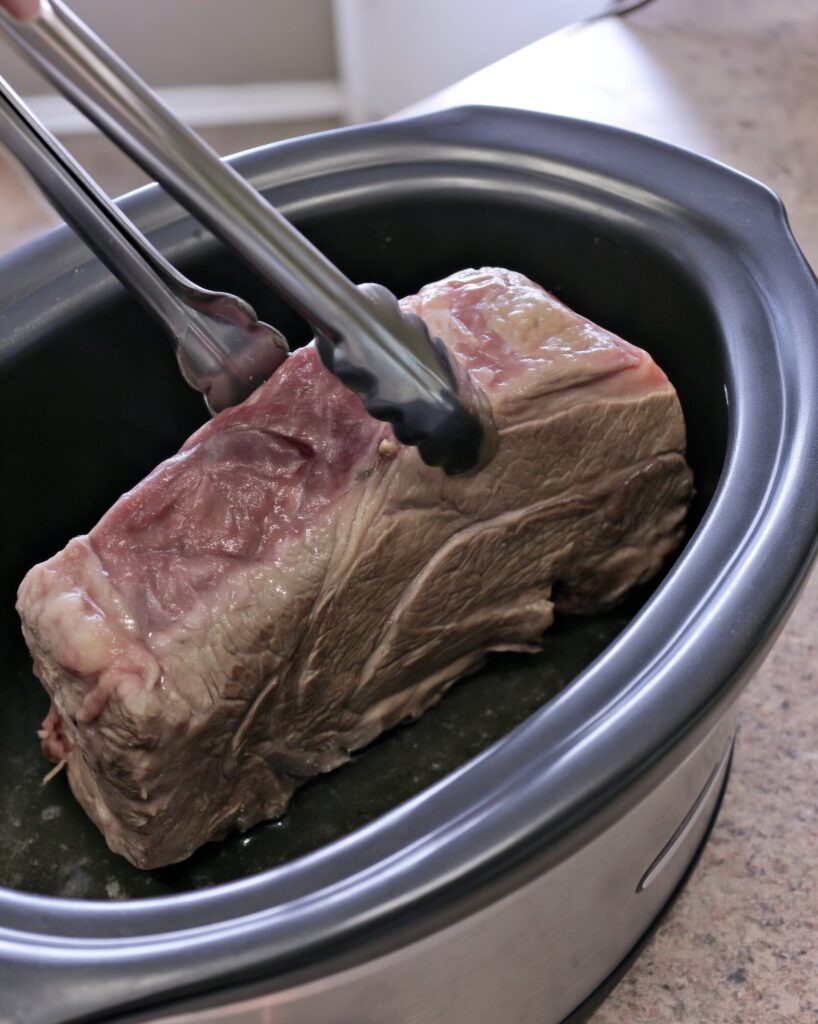 Metal tongs lifting a chuck roast in a black oval slow cooker insert on a kitchen counter