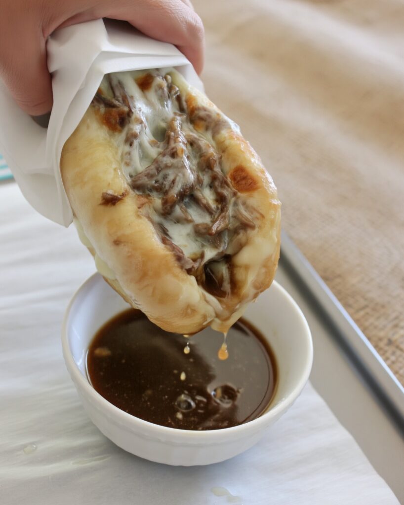 A French dip sandwich wrapped in parchment being dipped into a white bowl of dark au jus, with drips falling back into the bowl