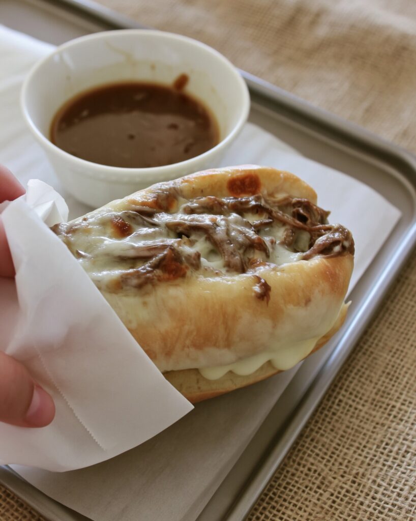 A shredded beef and provolone French dip sandwich held in a parchment wrap over a sheet pan, with a white bowl of au jus behind it