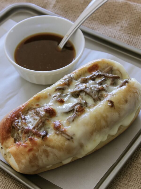 A hoagie roll filled with shredded beef and melted provolone on a parchment-lined sheet pan, with a spoon resting in a white bowl of au jus behind it