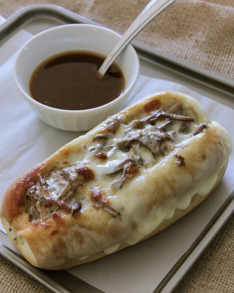A hoagie roll filled with shredded beef and melted provolone on a parchment-lined sheet pan, with a spoon resting in a white bowl of au jus behind it