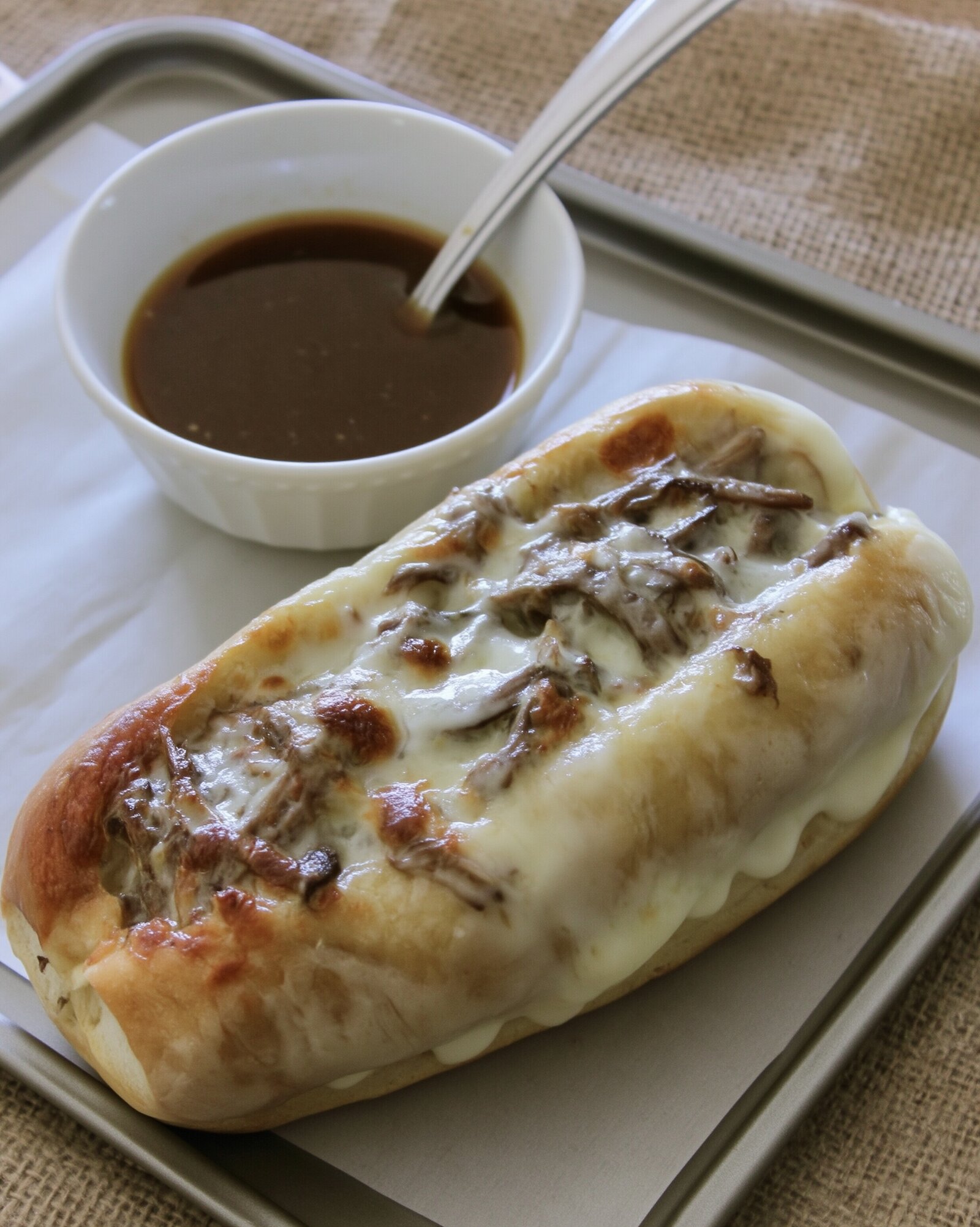 A hoagie roll filled with shredded beef and melted provolone on a parchment-lined sheet pan, with a spoon resting in a white bowl of au jus behind it