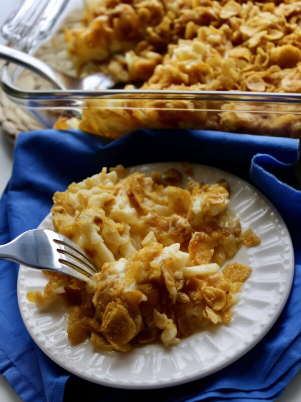 A serving of cheesy hashbrown casserole with crushed cornflake topping on a white fluted plate with a fork, on a blue napkin, the glass baking dish behind