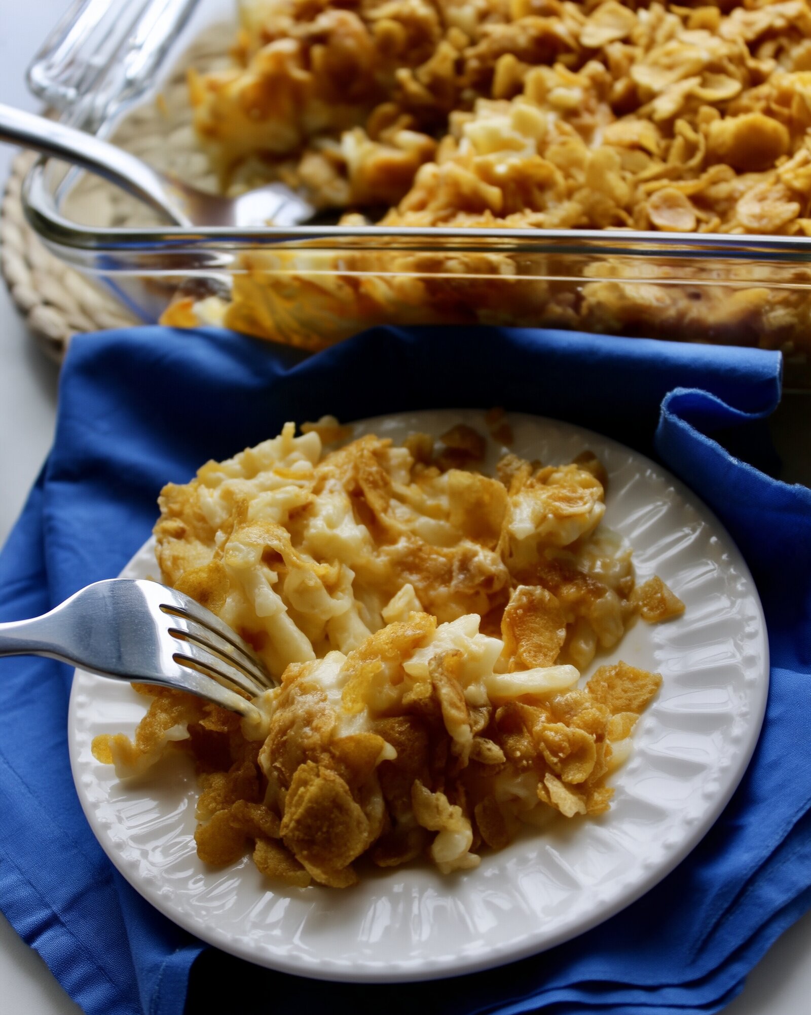 A serving of cheesy hashbrown casserole with crushed cornflake topping on a white fluted plate with a fork, on a blue napkin, the glass baking dish behind