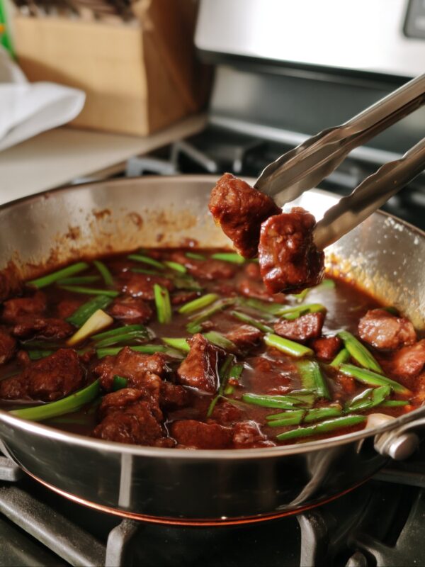 Metal tongs lifting pieces of saucy Mongolian beef out of a stainless steel pan with long-cut green onions on the stovetop