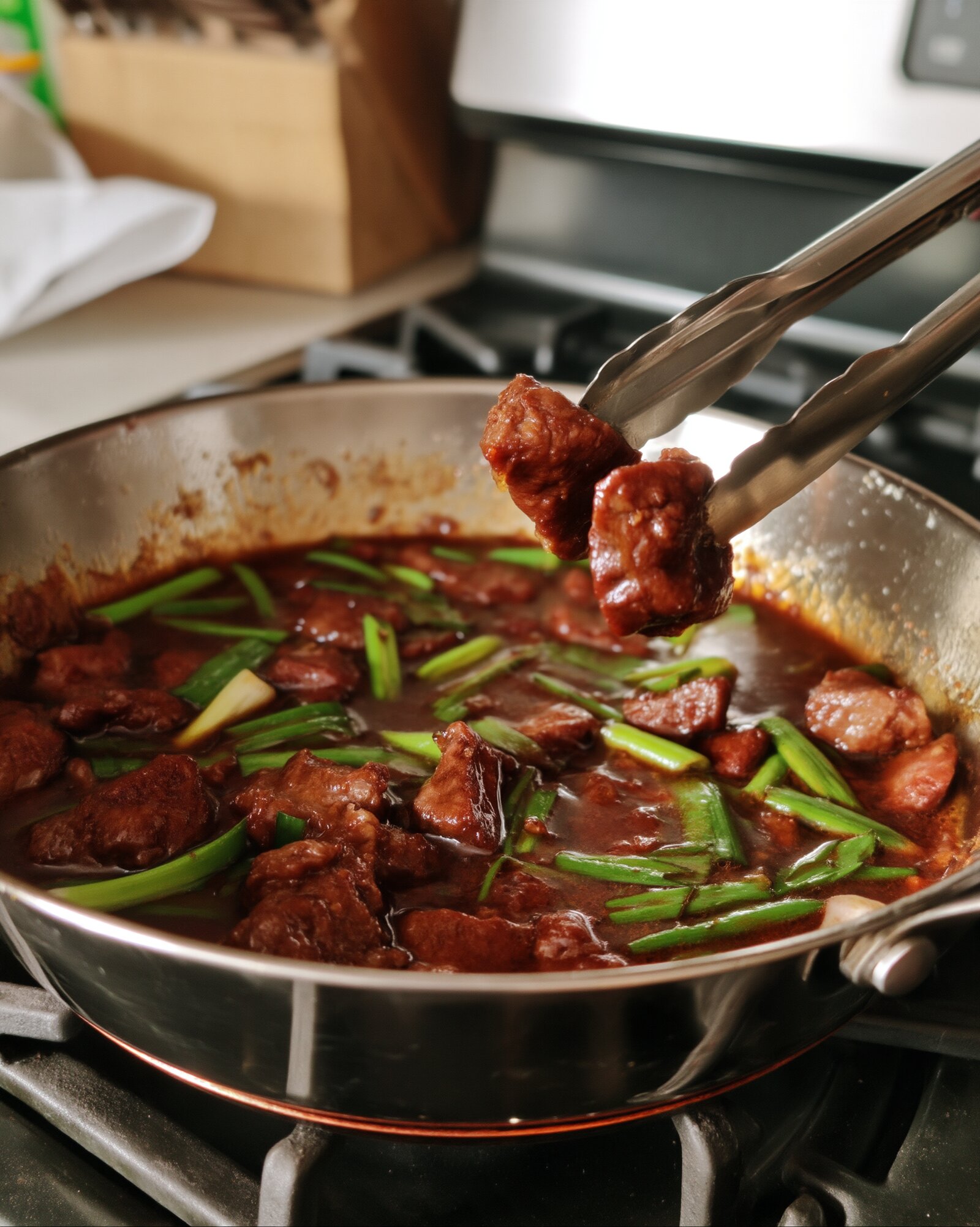 Metal tongs lifting pieces of saucy Mongolian beef out of a stainless steel pan with long-cut green onions on the stovetop