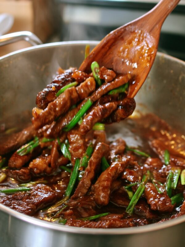 A wooden spoon tossing thin strips of crispy glazed flank steak with sliced green onions in a stainless steel skillet