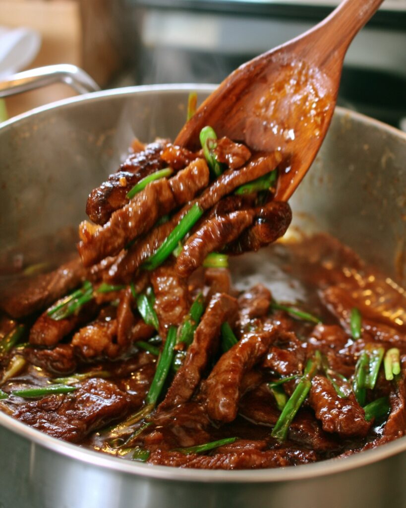 A wooden spoon tossing thin strips of crispy glazed flank steak with sliced green onions in a stainless steel skillet