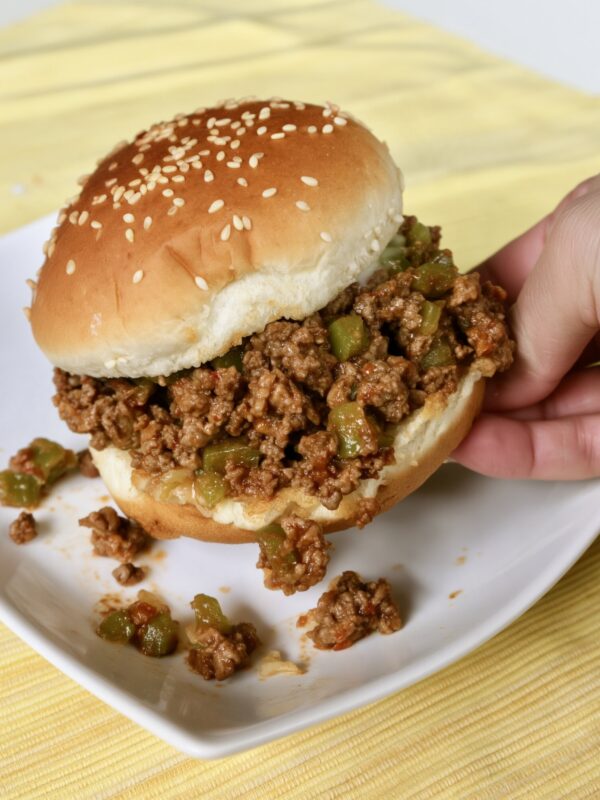 A sesame seed bun sloppy joe with ground beef and chunky green pepper being lifted from a white plate, some filling spilled on the plate, yellow striped placemat