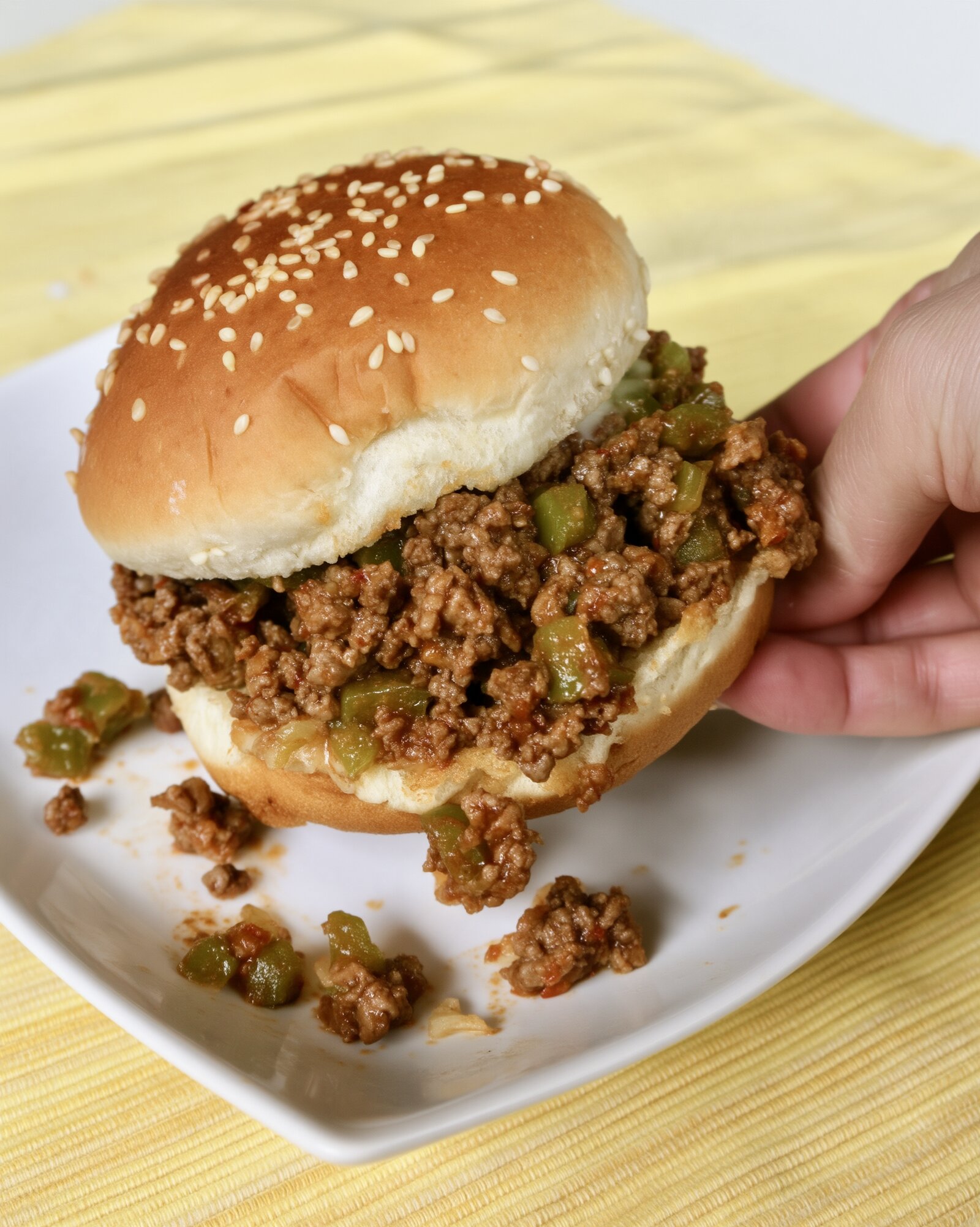 A sesame seed bun sloppy joe with ground beef and chunky green pepper being lifted from a white plate, some filling spilled on the plate, yellow striped placemat