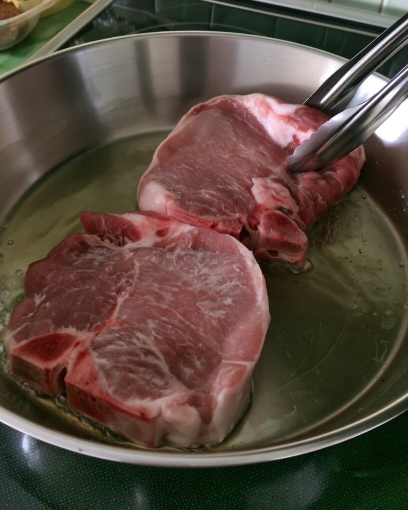 Two raw bone-in pork chops in a stainless steel skillet with a little oil, metal tongs lifting one chop