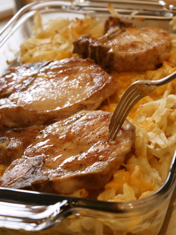 Seared bone-in pork chops resting on cheesy shredded hashbrowns in a glass baking dish, a fork lifting the edge of one chop