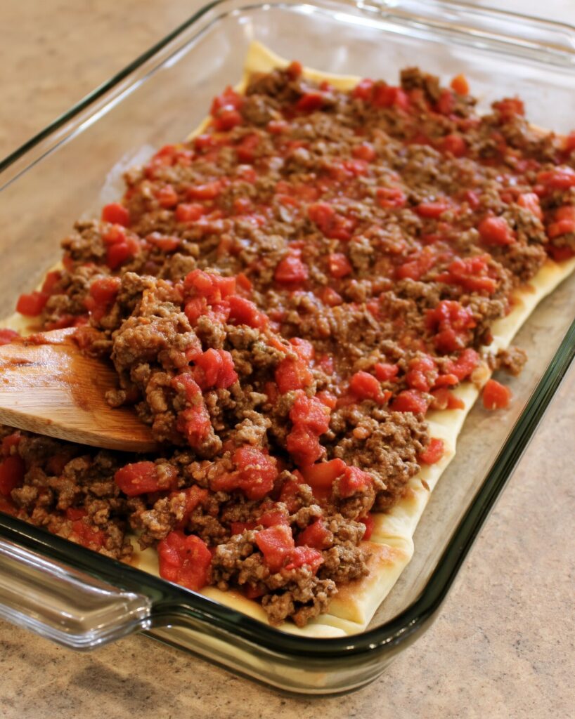 Crumbled seasoned ground beef and drained diced tomatoes piled on a wooden spoon in a glass baking dish lined with crescent roll dough