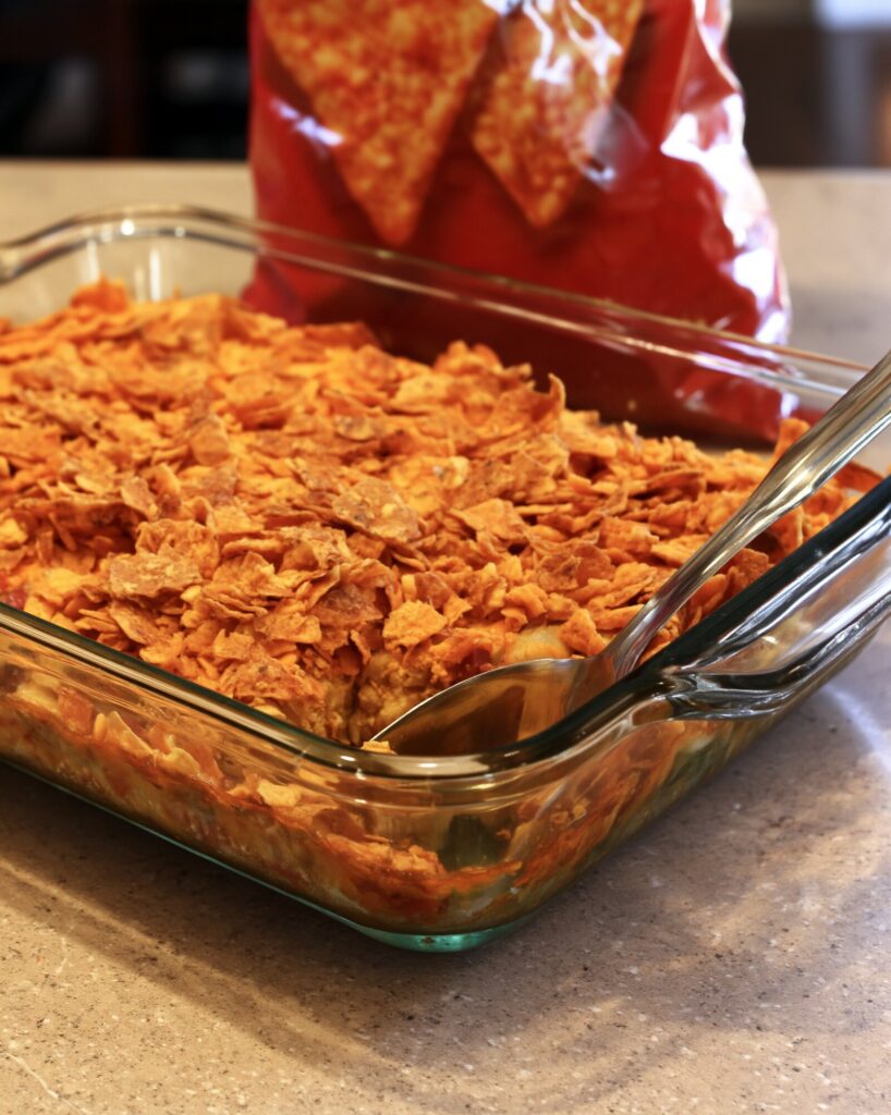 Taco crescent bake seen from countertop level through the side of a glass baking dish, with a serving spoon in the corner and crushed nacho cheese chips covering the top