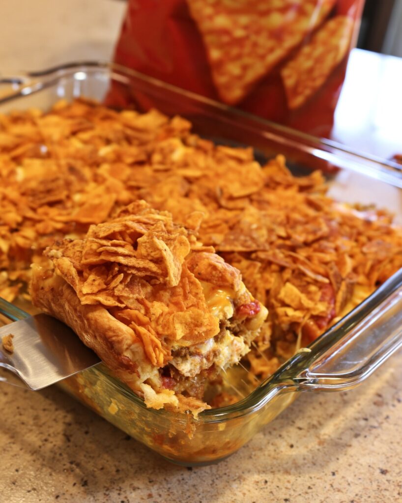 A serving of taco crescent bake lifted from the corner of a glass baking dish on a speckled countertop, crushed nacho cheese chips piled on top and the soft crescent crust underneath