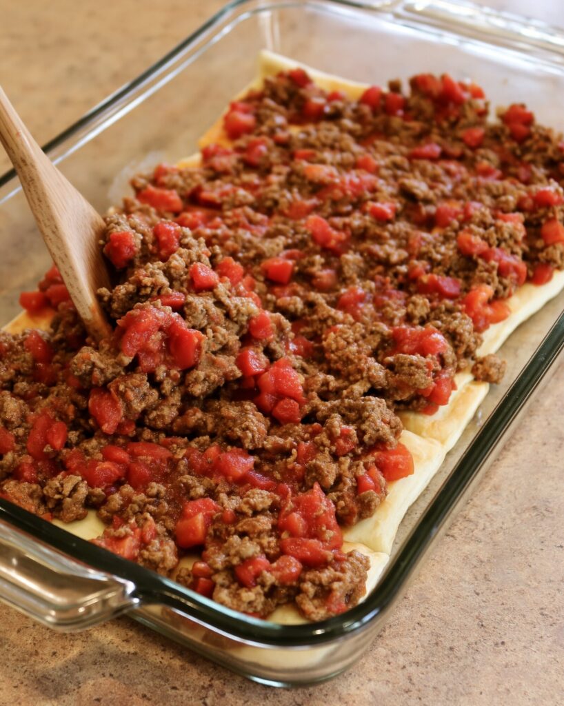 A wooden spoon spreading seasoned ground beef and diced tomatoes over crescent roll dough in a glass 9x13 baking dish