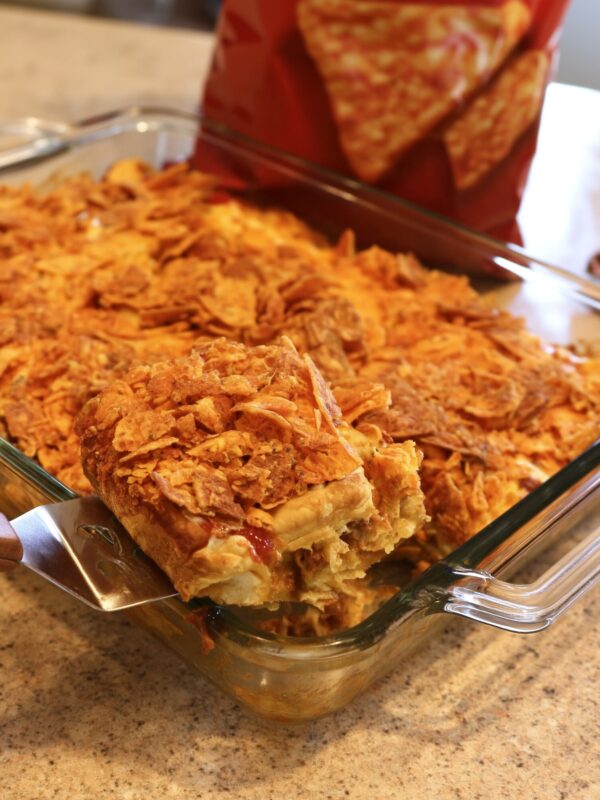 A metal spatula lifting a square of taco crescent bake out of a glass 9x13 dish, showing the crescent crust, seasoned beef and a thick crushed nacho cheese chip topping, with a red chip bag behind it