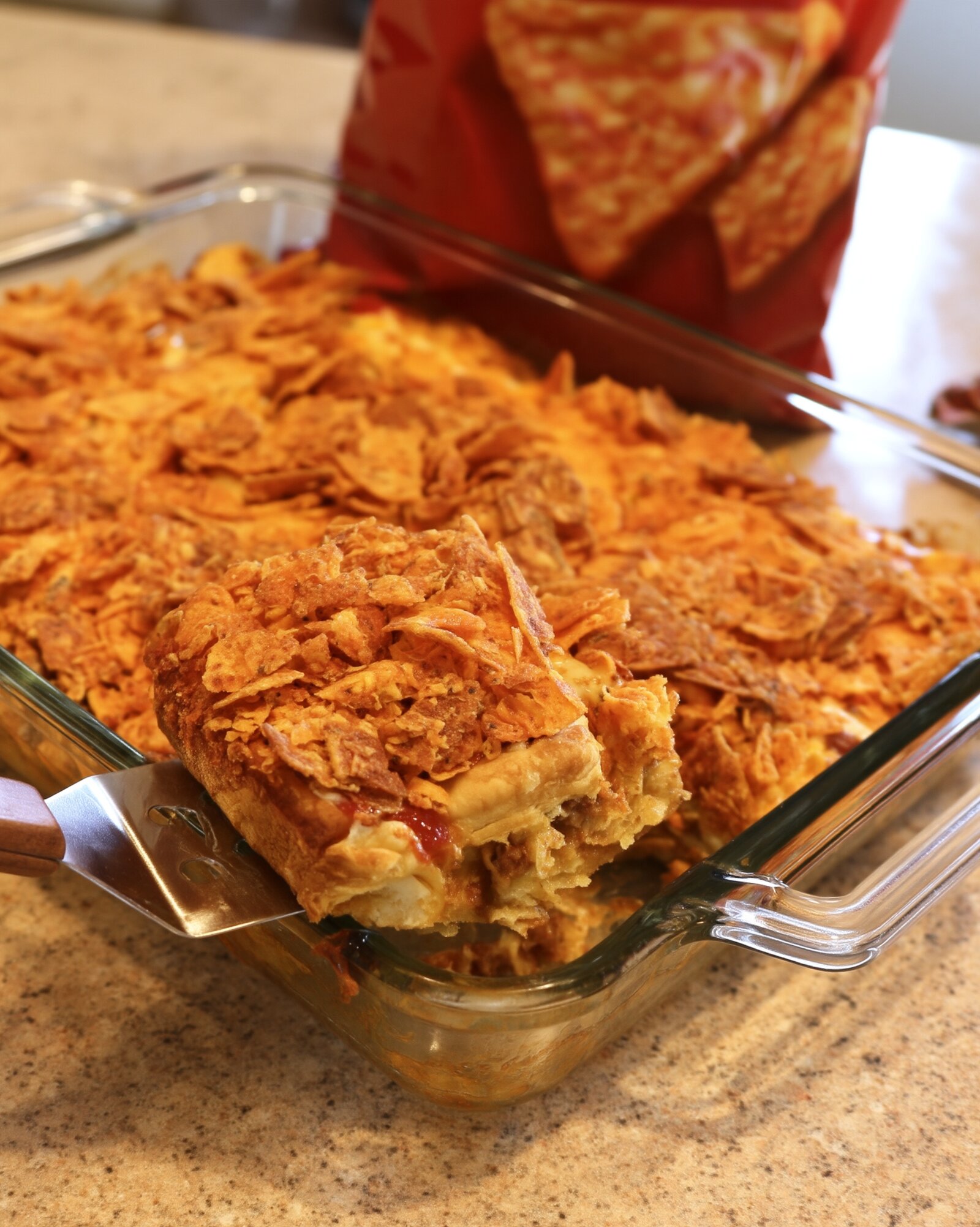 A metal spatula lifting a square of taco crescent bake out of a glass 9x13 dish, showing the crescent crust, seasoned beef and a thick crushed nacho cheese chip topping, with a red chip bag behind it